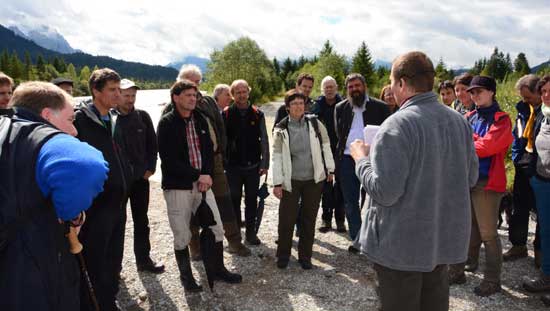 Bernhard Schaipp in der Bildmitte erläutert den Teilnehmern und anderen Referenten die geplanten Maßnahmen. Im Hintergrund die Isar mit Auwald und Bergkulisse.