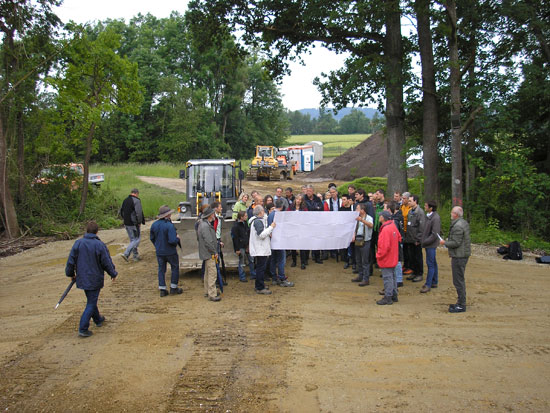 Baustelle mit Radlader; ein großer Plan wird von zwei Teilnehmern gehalten, die übrigen Teilnehmer stehen im Halbkreis dahinter.