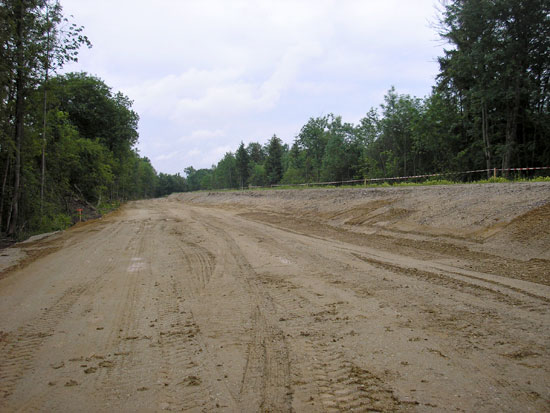 Vegetationslose, breite Baustraße und rechts angrenzend alter Deich, dessen Vegetation entnommen wurde. Rechts und links jeweils Wald.