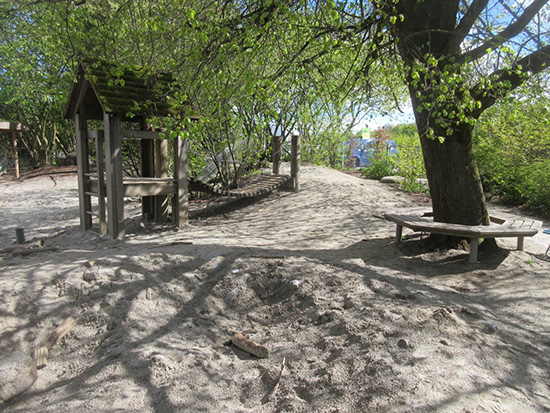 Neben einer großen Sandmulde steht ein Baum, der von einer Sitzbank umgeben ist, daneben ein kleines Spielhaus mit Kettenbrücke.