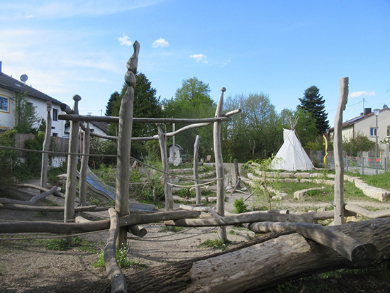 Eine große Spielfläche, bestehend aus mehreren dünnen Holzstämmen, die vertikal und horizontal zu einem Klettergerüst verbunden sind. Dahinter sind eine Rutsche und ein Tipi zu sehen.