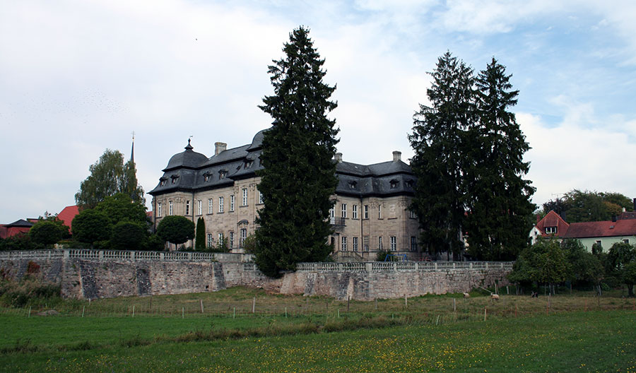 Das Amtsschloss Burgwindheim liegt etwas erhoben über der umgebenden Wiesenlandschaft, gesäumt von einigen Nadelbäumen.